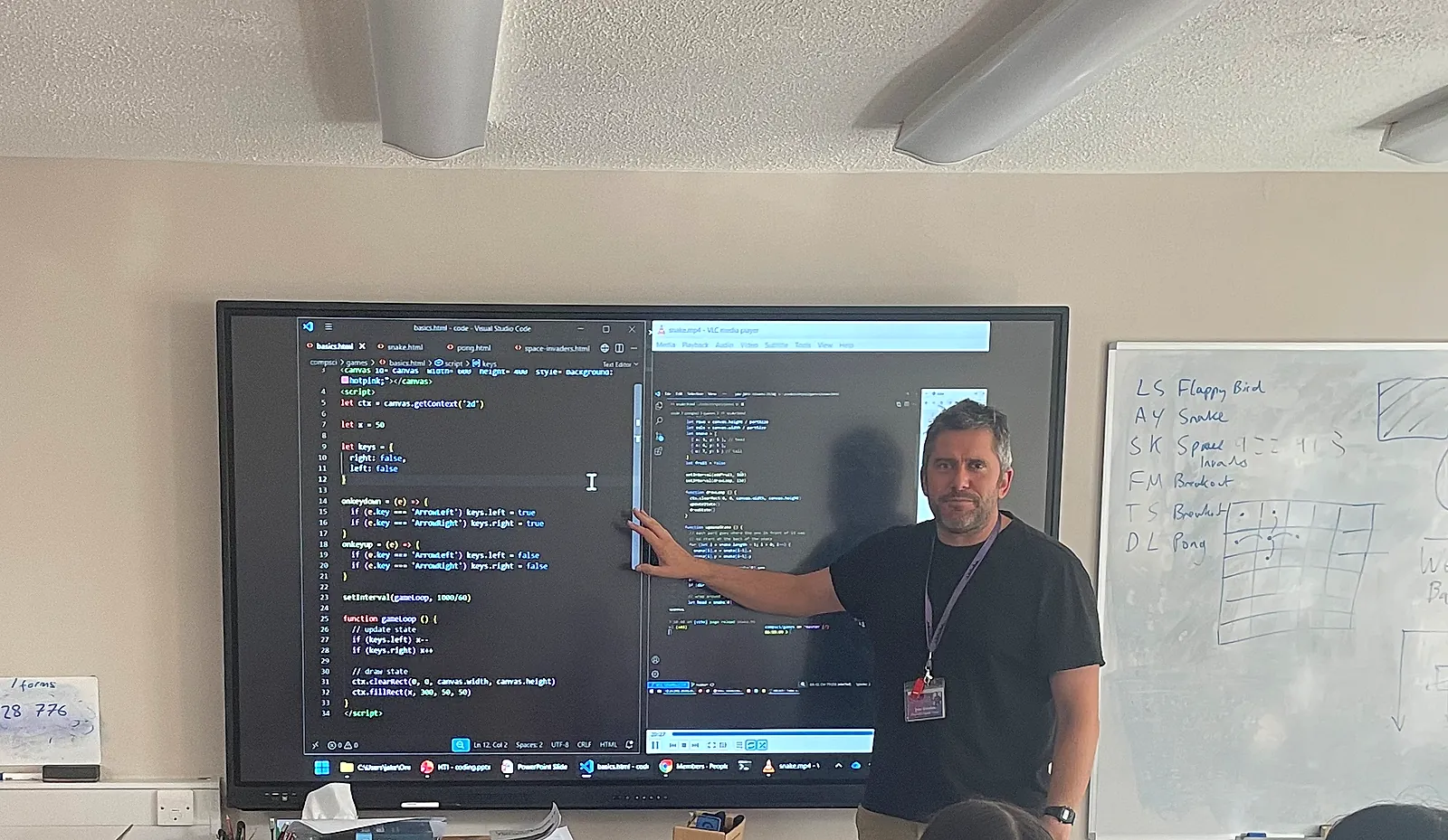 Jake Gordon at the front of a classroom, pointing at code for a browser game on a large screen