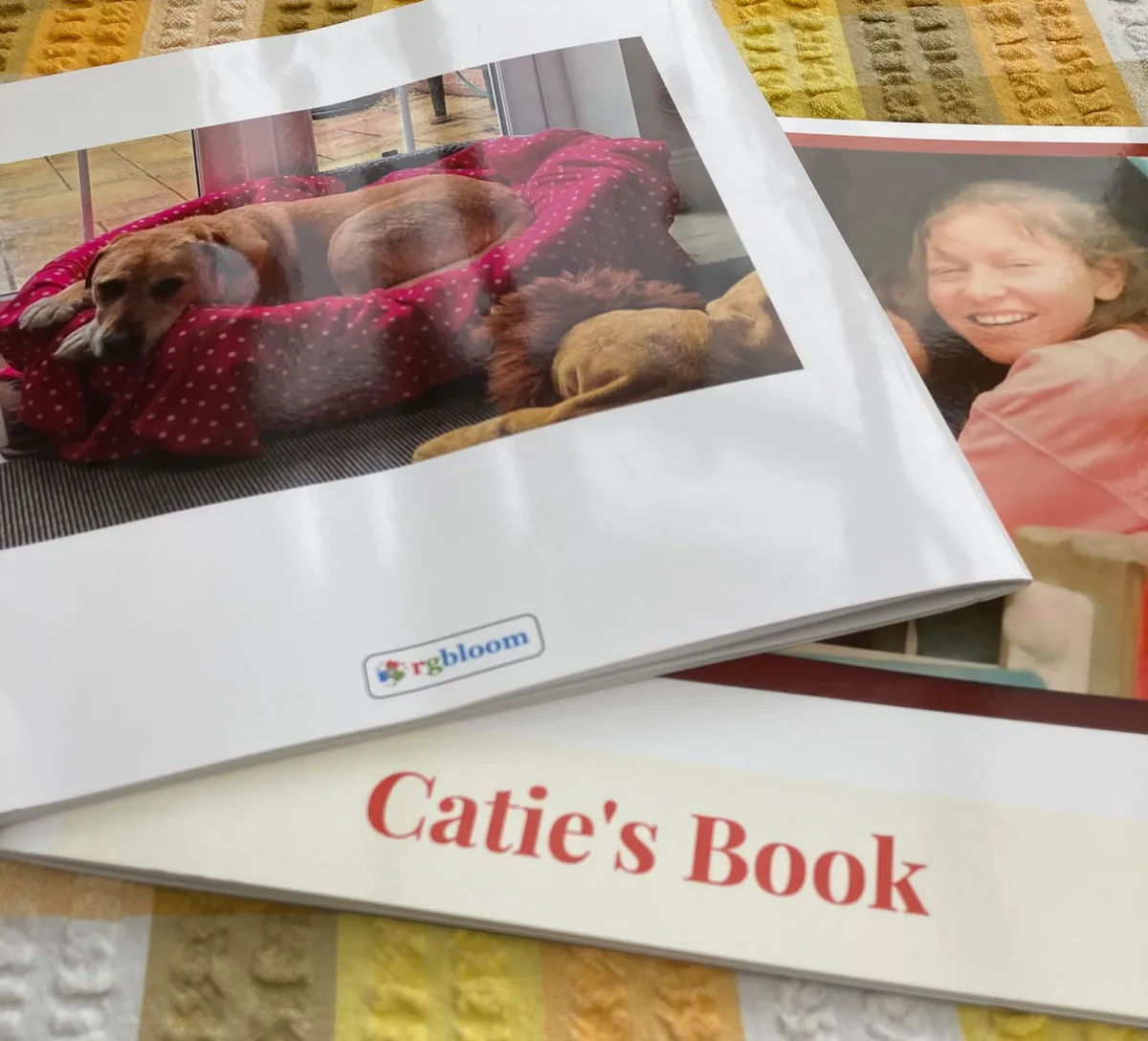 Two printed photo books on a yellow striped cloth: one open at a dog curled up asleep in a red spotted bed, the other with a smiling woman on the cover, titled Catie's Book