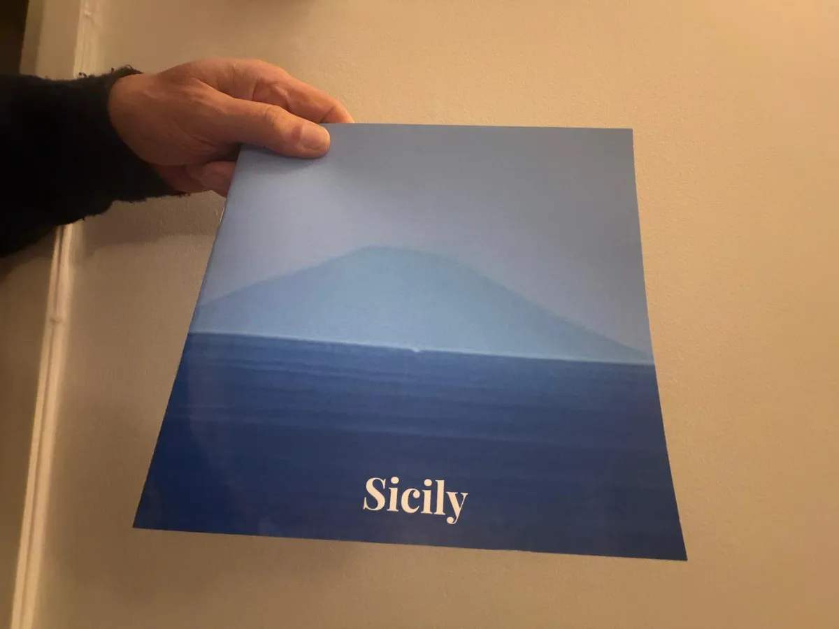 A hand holding up a printed photo book, its blue cover a misty mountain above the sea, titled Sicily
